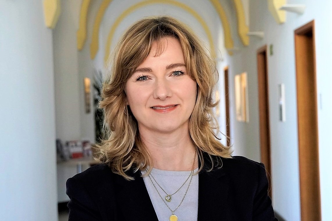 Eine Frau mit schulterlangen Haaren in einem historischen Flur, der sanft erleuchtet ist. 