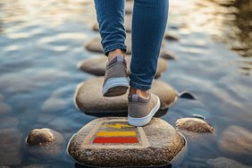 Zwei Beine mit Füßen in Turnschuhen balancieren auf großen Steinen im Wasser, wobei der erste Stein mit einer gemalten Energielade-Anzeige bemalt ist. 
