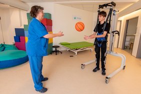Eine Therapeutin spielt mit einem Jugendlichen Ball, der von einem großen Gehtrainer gestützt wird