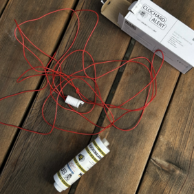Nahaufnahme des Clochard Alert, einer kleinen, etwa acht Zentimeter langen elektronischen Alarmanlage mit einem roten Sicherungskabel. Das Gerät ist so konzipiert, dass obdachlose Menschen ihre persönlichen Sachen damit sichern können. Wird das Kabel gewaltsam entfernt, löst das Gerät einen Alarm aus.